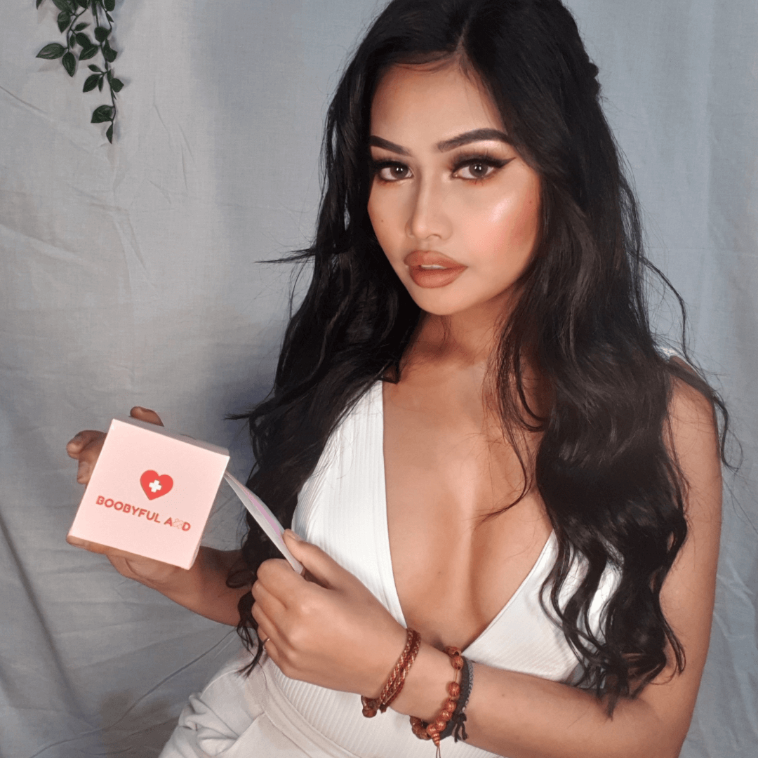 girl with black curly hair in white background holding boobyful aid box pulling clear tape from the slit in a white low cut v bodysuit using clear tape