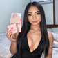 girl with long black hair sitting on bed holding boobyful aid box and nipple covers in low v bodysuit wearing clear boob tape for lift
